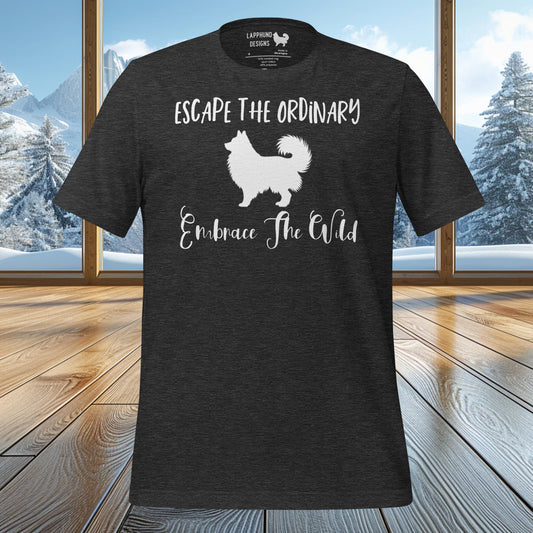 A black t-shirt featuring a silhouette of a Finnish Lapphund with the phrases 'Escape The Ordinary' and 'Embrace The Wild' in bold white text. The t-shirt is displayed in front of a cozy cabin scene with snowy mountain peaks visible through the windows, reinforcing the outdoor adventure theme.