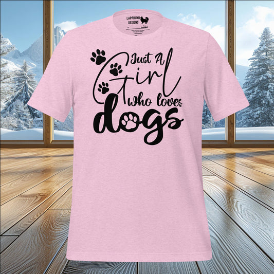 A pink t-shirt with the phrase 'Just a Girl Who Loves Dogs' in bold black text, accented with paw prints. The t-shirt design includes a playful paw print within the word 'dogs' for added charm. Displayed against a cozy cabin setting with snow-covered mountains visible through the windows.